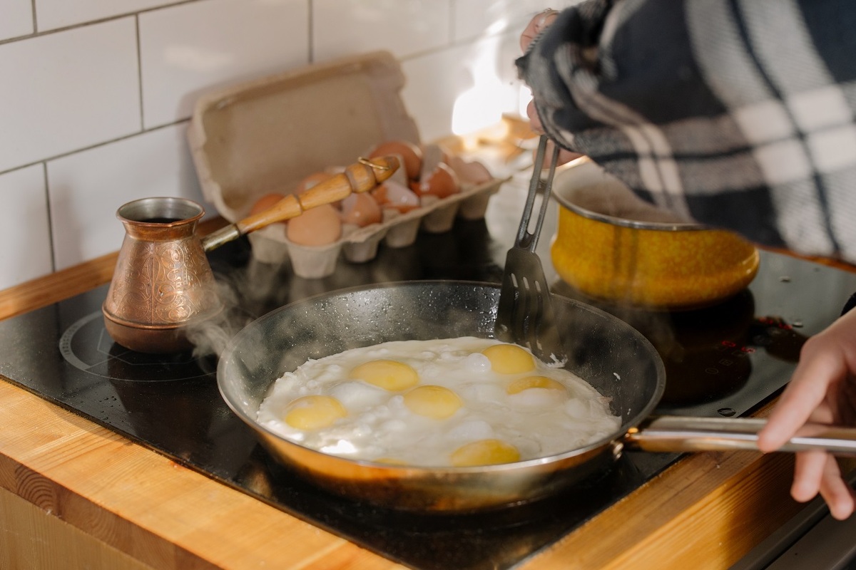 Imagem de uma frigideira no fogão com ovos sendo fritos, ao fundo uma caixa de ovos aberta
