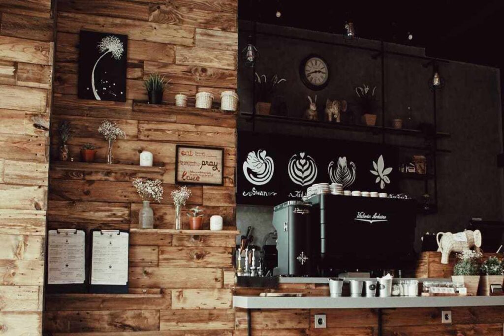Cafeteria com tema rústico onde a parede de madeira segura quadros e o foco da imagem é uma cafeteira preta e por trás dela uma prateleira com xícaras e copos