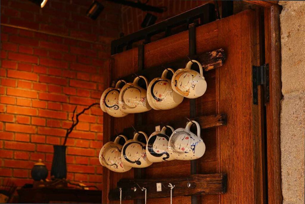 Imagem com foco na prateleira com canecas de café com vários desenhos diferentes. Prateleira de madeira em pallet em parede rústica sem acabamento.