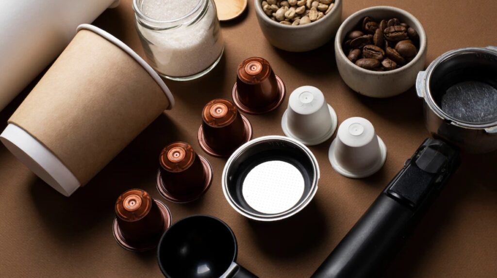 Cápsulas de café sobre mesa e bows com grãos, além de xícaras e copos e um pote de açúcar