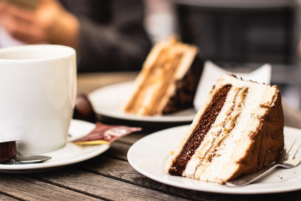 Mesa com pessoa sentada ao fundo e sobre ela dois pedaços de bolo em pires brancos e uma xícara grande de café ao lado.
