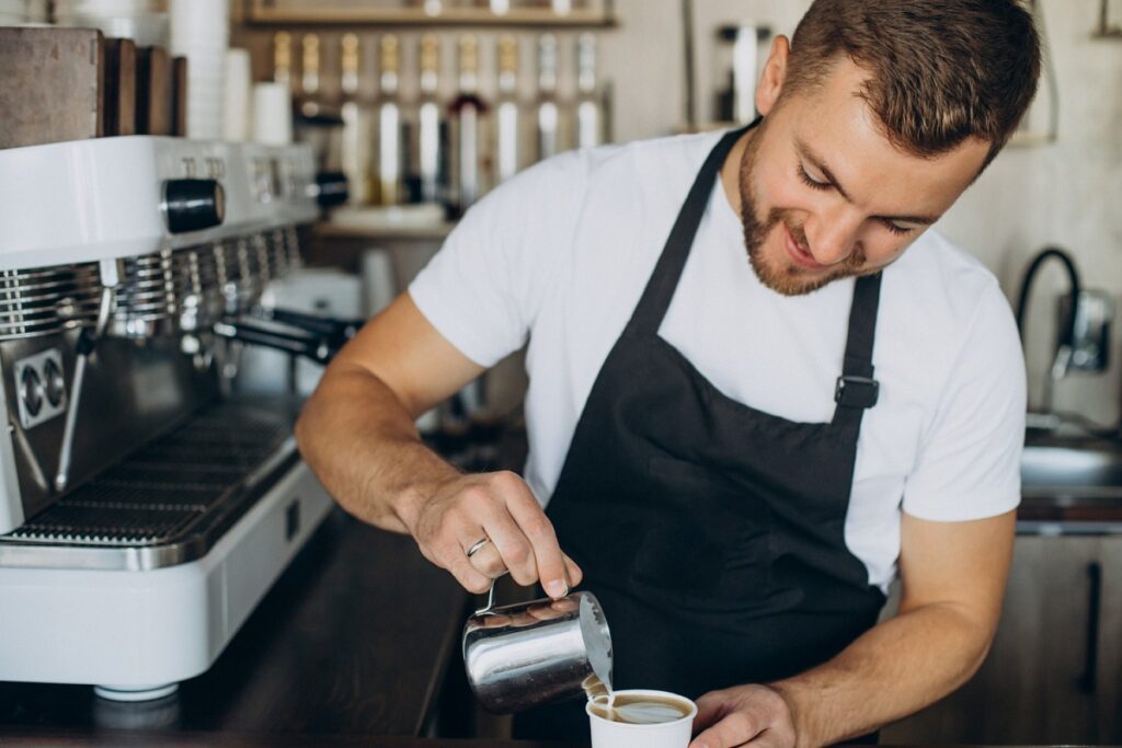 Barista preparando café espresso com leite em cozinha de cafeteria com máquinas ao seu lado e fundo em desfoque.