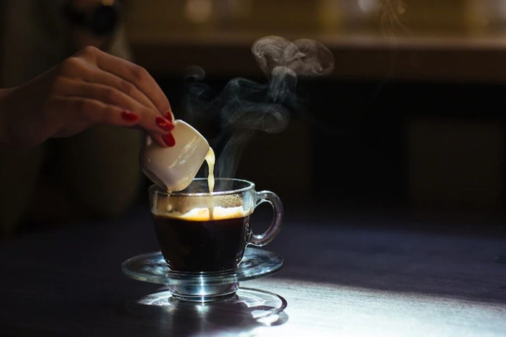 Mulher derrama leite em café feito por capsulas Bicafé em xícara grande sobre pires transparente em cima de mesa.