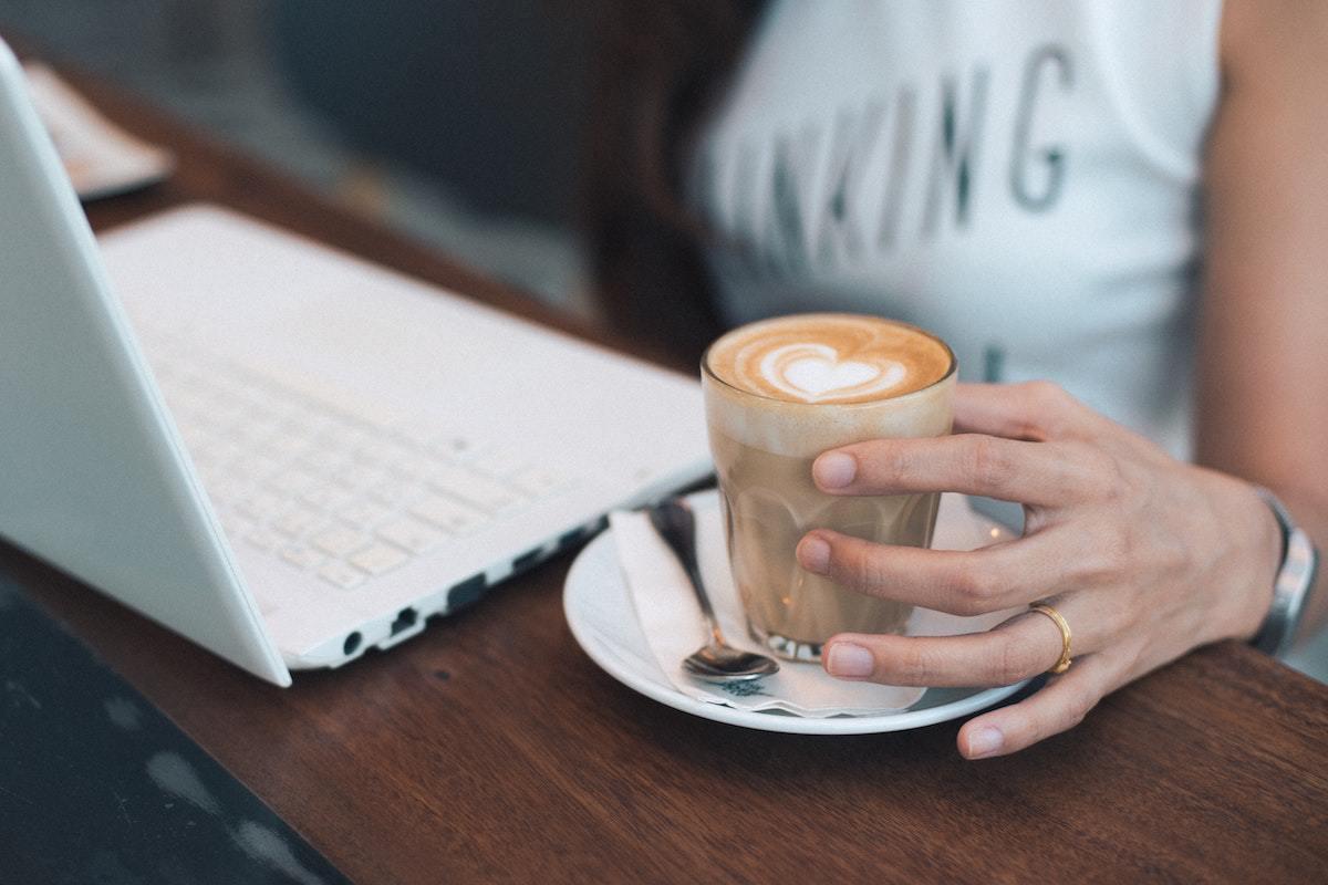 Mulher usando notebook branco com caneca de café espresso ao seu lado esquerdo, que ela segura com mão que tem relógio prata no pulso e aliança dourada no dedo anelar