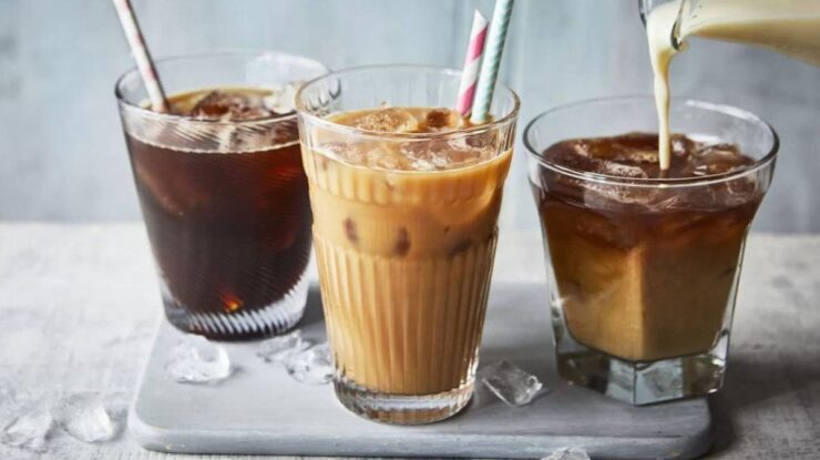 Três copos diferentes de bebidas feitas com café enquanto pessoa derrama leite dentro de um deles, que estão em cima de base de madeira cinzenta em mesa com pano claro que a cobre