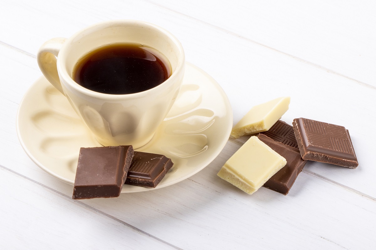 Xícara de café sobre pires na cor creme com bebida preta em seu interior e tabletes quadrados de chocolate branco e preto espalhados ao redor