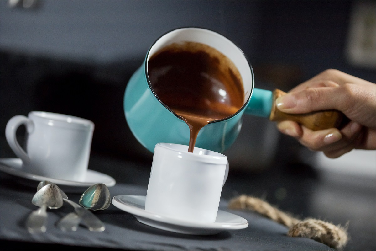 Duas xícaras brancas de café sobre pires em cima de mesa de cor escura enquanto pessoa derrama chocolate quente dentro de uma delas