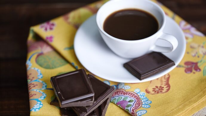 Xícara de café preto sobre pires branco em cima de pano de mesa dobrado com alguns pedaços de barra de chocolate espalhados ao lado