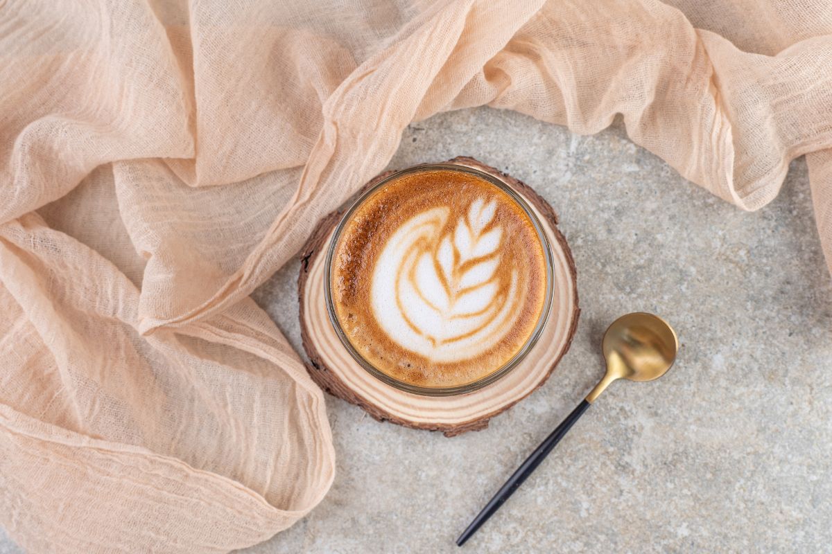 Café espresso com leite formando desenho dentro de xícara sobre suporte redondo de madeira em cima de mesa de pedra com colher dourada ao lado e lençol fino de cor salmão engelhado do outro