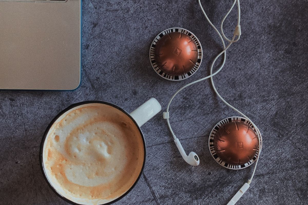 Café espresso em caneca preta e branca sobre mesa cinzenta ao lado de notebook, fones de ouvido e duas cápsulas de café em cor metálica laranja