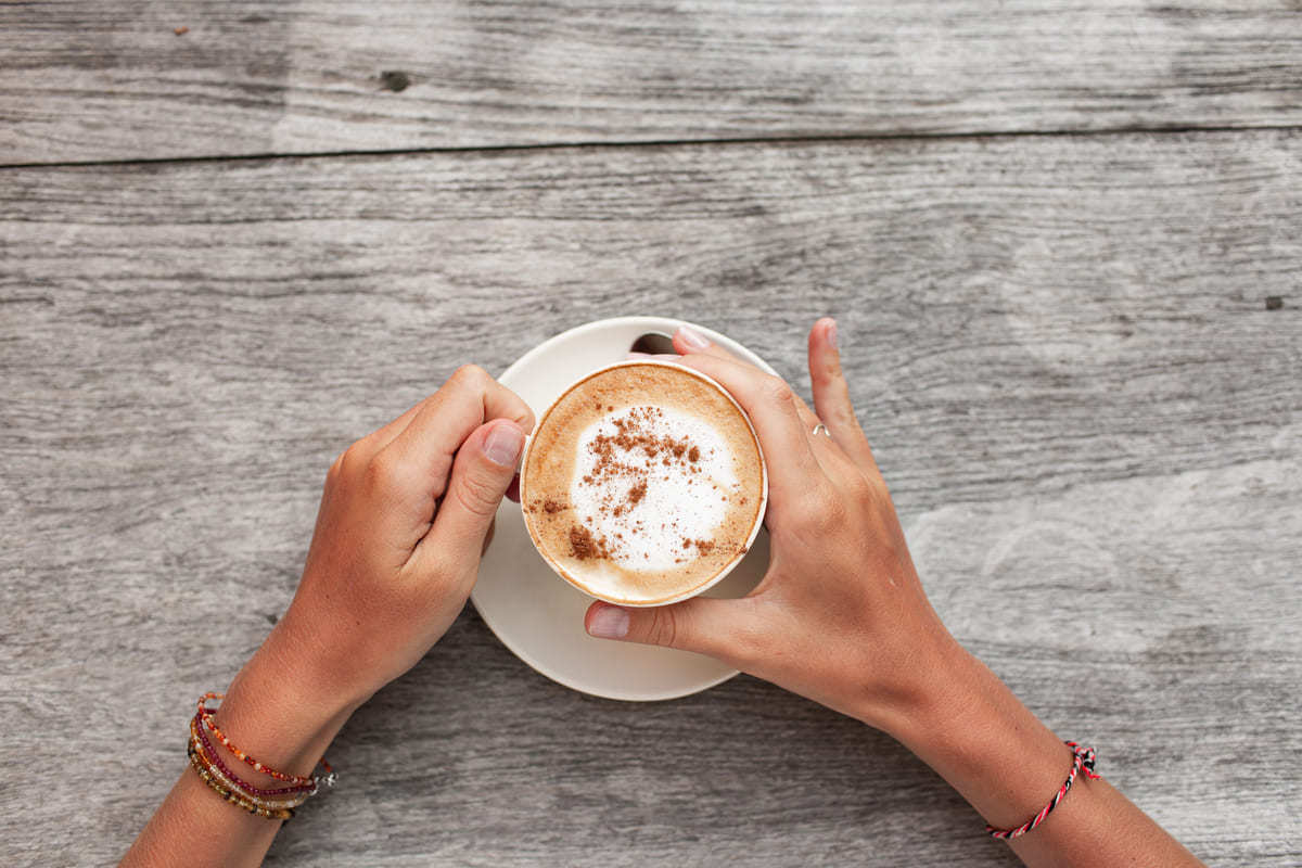 Mãos femininas apoiam xícara grande de café expresso com leite sobre pires branco em cima de mesa cinza