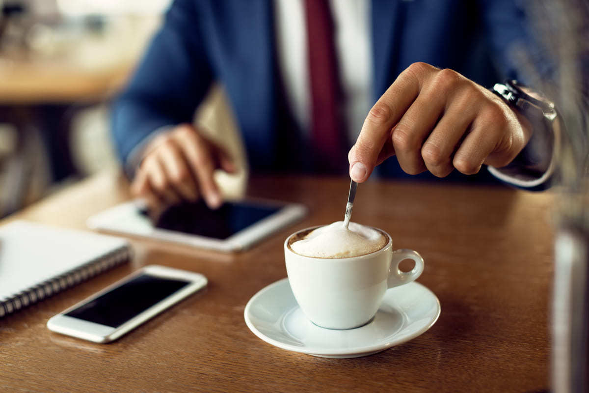 Homem de paletó azul sentado diante de mesa de madeira escura mexendo seu café em xícara grande branca sobre pires branco com uma agenda e um tablet, além de um smartphone, ao lado da mesma