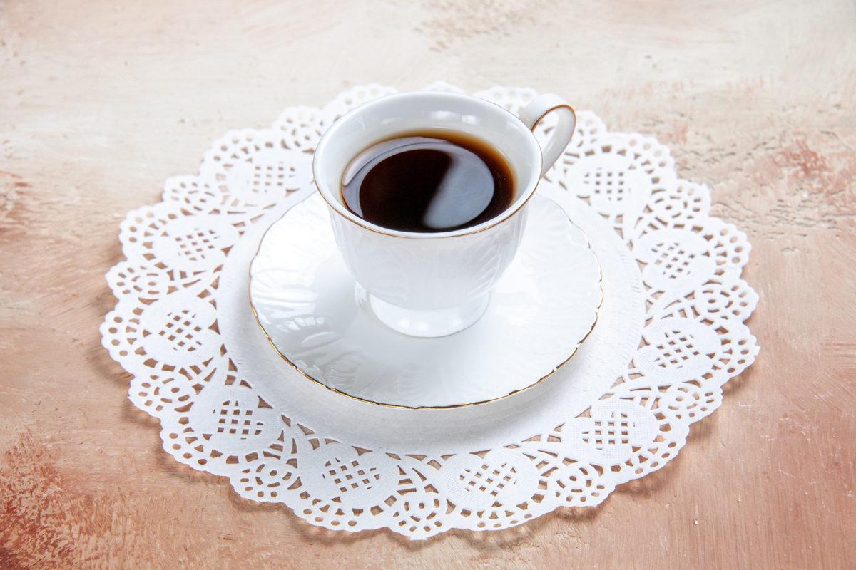 Xícara pequena com café preto sobre pires e prato branco com bordas em decoração análoga à renda em mesa de madeira