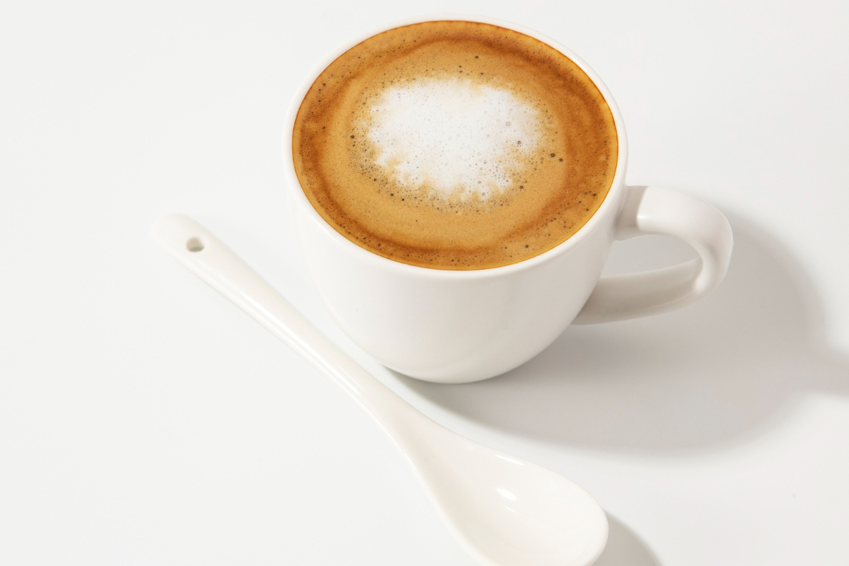 Xícara branca média com café espresso com leite em cima de superfície branca e ao lado de colher grande da mesma cor