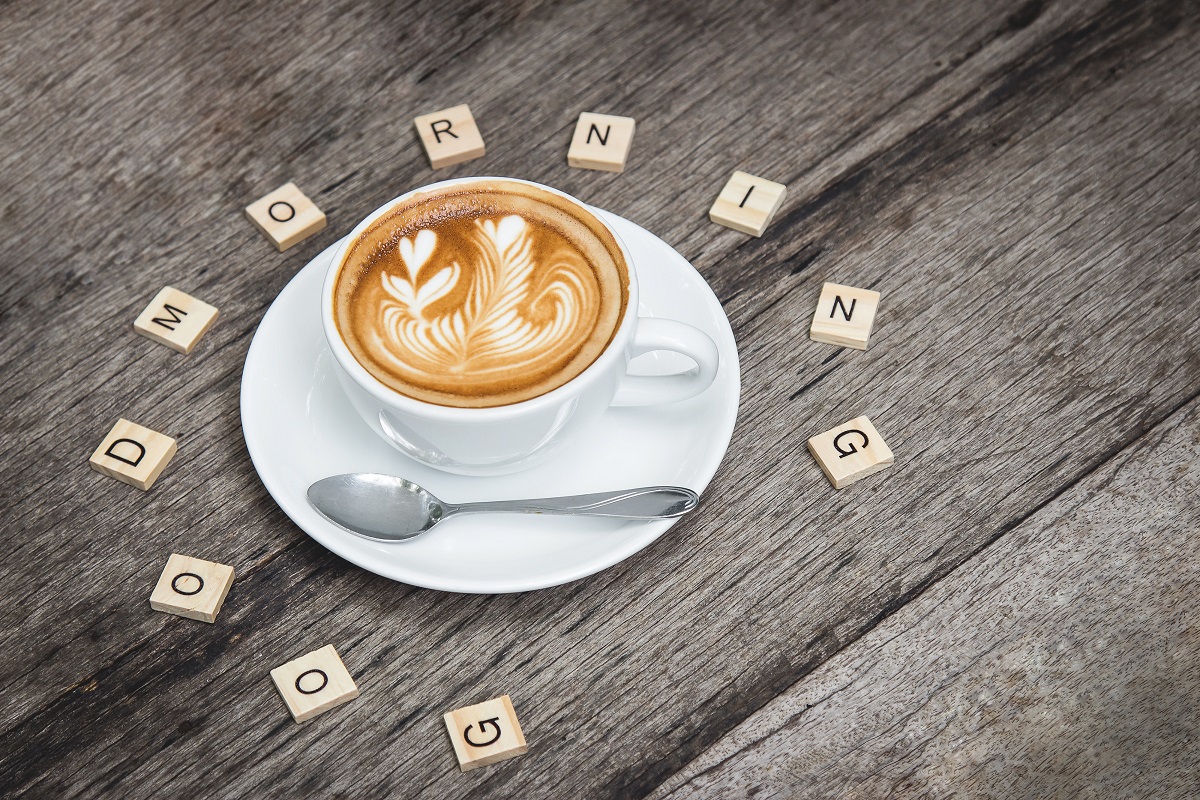 Xícara branca de café com leite formando desenho na sua superfície sobre pires branco em cima de mesa cinzenta de madeira com letras gravadas em blocos de madeira ao redor da xícara formando a frase good morning