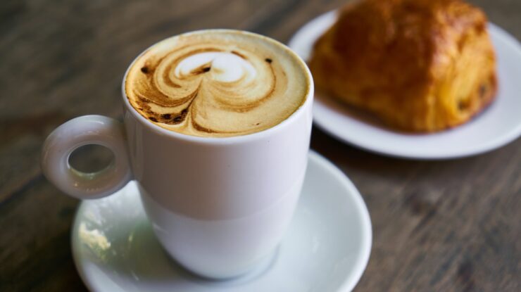 Foto de um dos Tipos de café da manhã com café espresso com leite em xícara comprida em cima de pires e com pão ao lado em desfoco sobre mesa de madeira