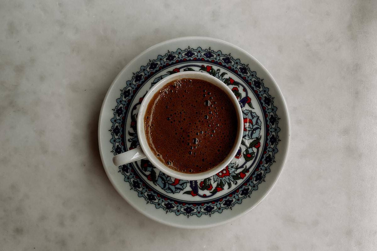 Foto tirada de cima de xícara com café preto espresso sobre pires decorado com mandala azul e amarela sobre mesa de pedra branca