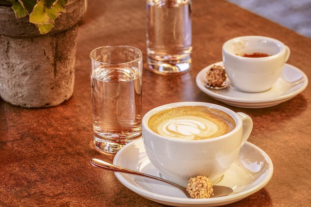 Xícara de café complexo ao lado de xícara menor sobre pires branco, com doce em colher e ao lado de copos de água sobre mesa de madeira em cafeteria