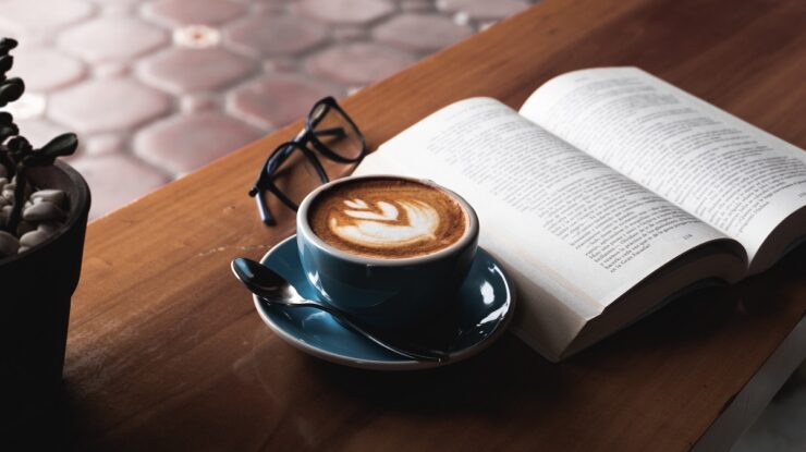 Livro aberto em mesa de madeira ao lado de xícara de café espresso e óculos dobrado em local de pouca luz