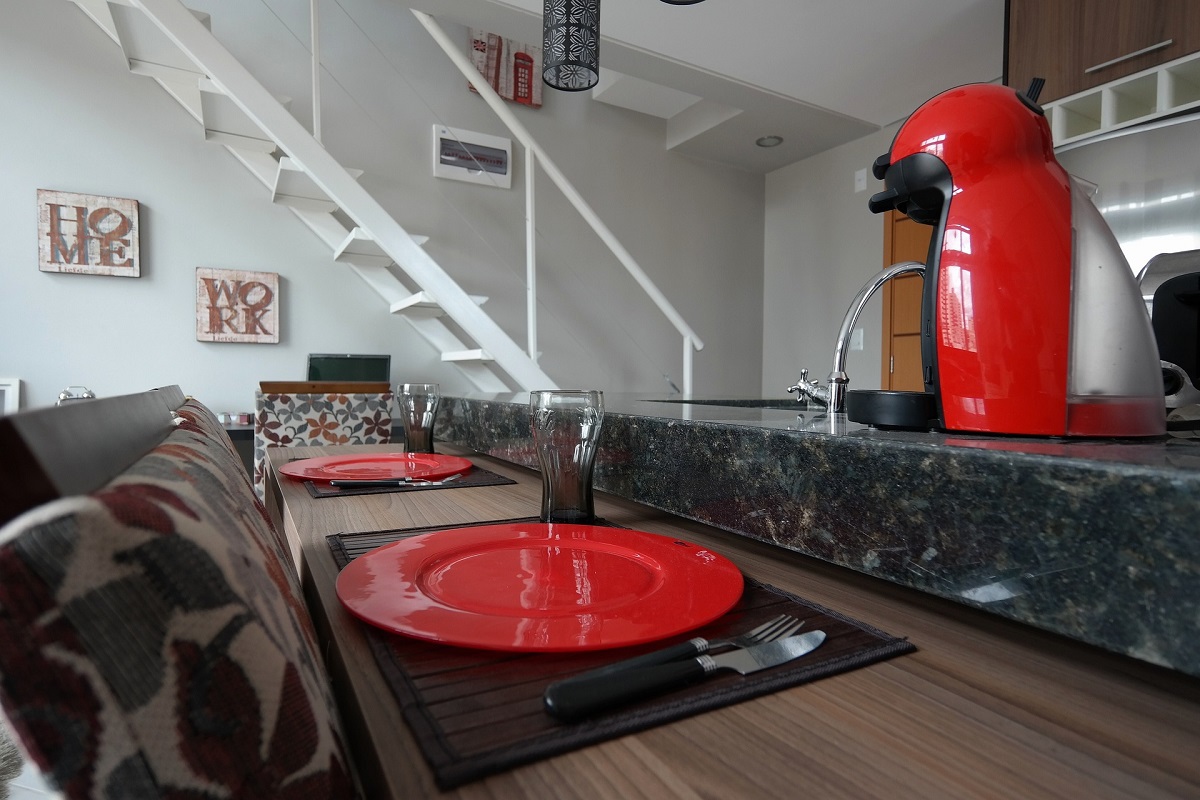 Balcão em cozinha de casa com uma das Melhores cafeteiras de cápsula sobre o mesmo e com dois pratos vermelhos diante de bancos e ao lado de copos de vidro transparente