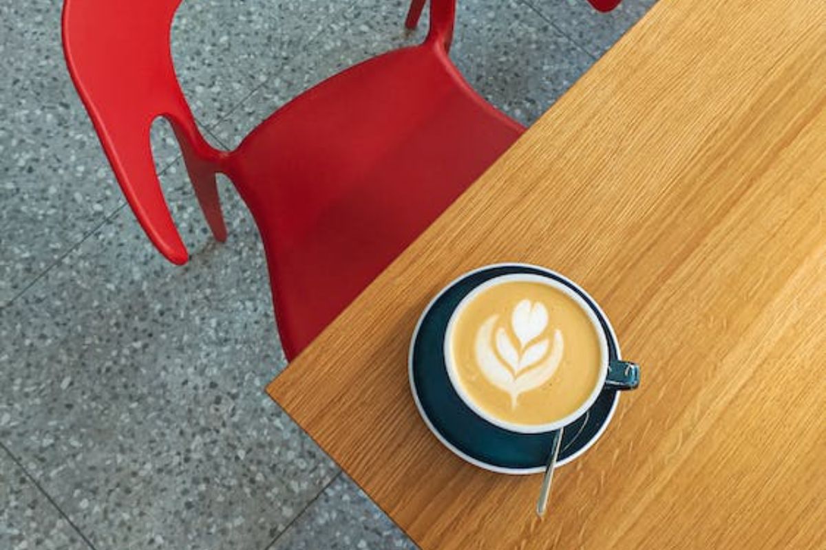 Foto tirada por Apaixonado por café da sua xícara de bebida espresso sobre mesa de madeira com cadeira vermelha diante da mesma em local com piso de pedras cinzentas