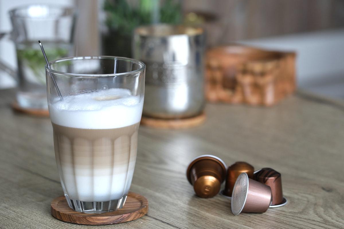 Café alemão em caneca sobre mesa de madeira com cápsulas à direta e objetos ao fundo em desfoco para preparo da bebida