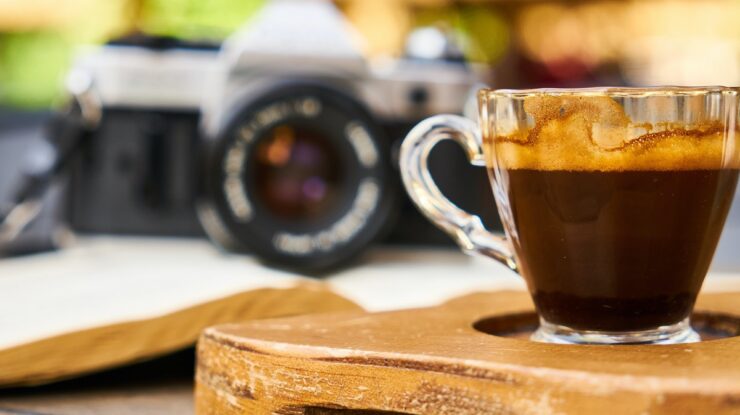Café no calor representado por xícara sobre suporte de madeira em cima de mesa com câmera semi profissional ao fundo em desfoco