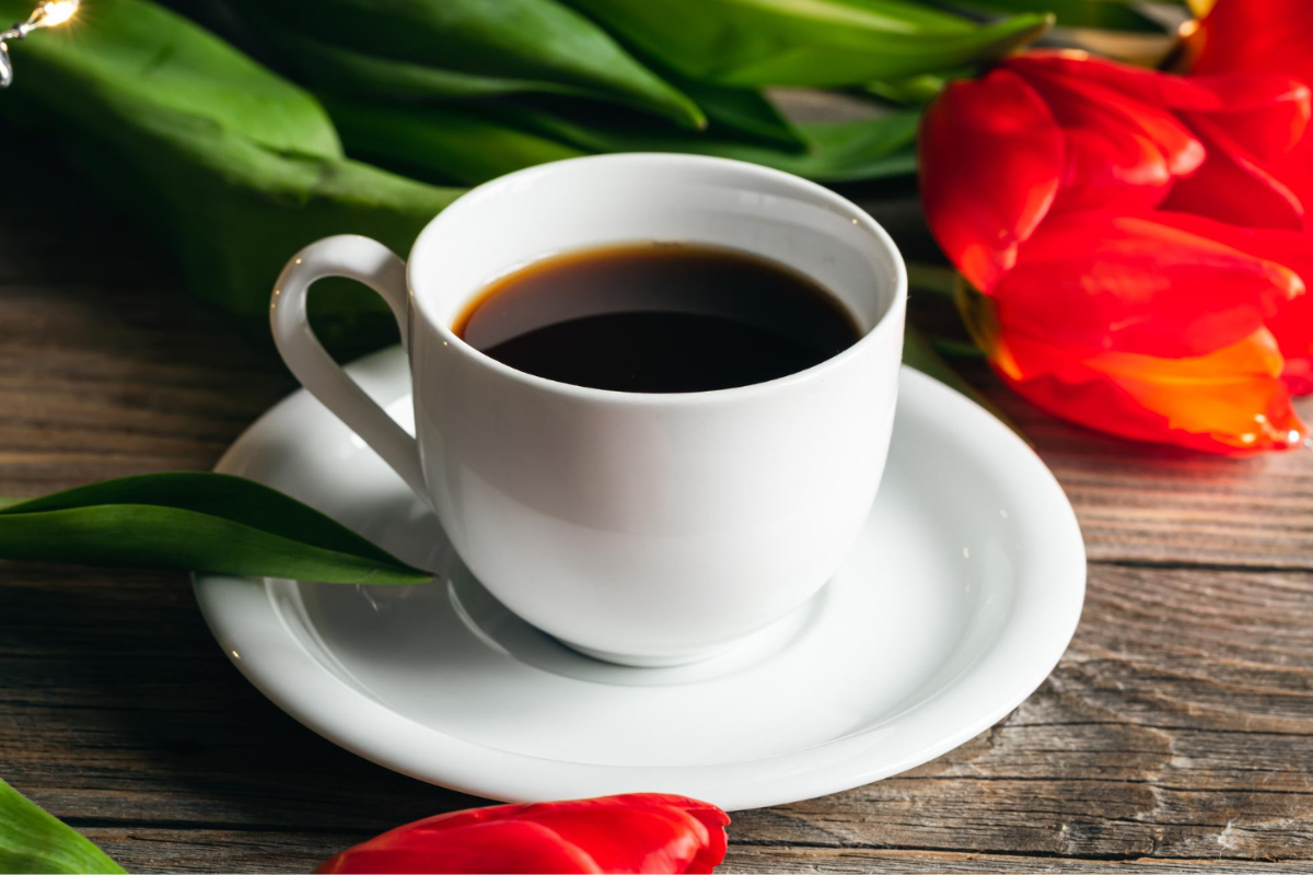 Café em xícara branca sobre pires de em mesa de madeira e ao lado de folhas e pimentas vermelhas
