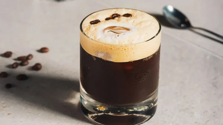 Close-up de um copo de carajillo, um coquetel espanhol cremoso de café e licor, com espuma clara, grãos de café decorativos e gelo visível, sobre uma superfície clara com grãos de café espalhados e uma colher ao lado.