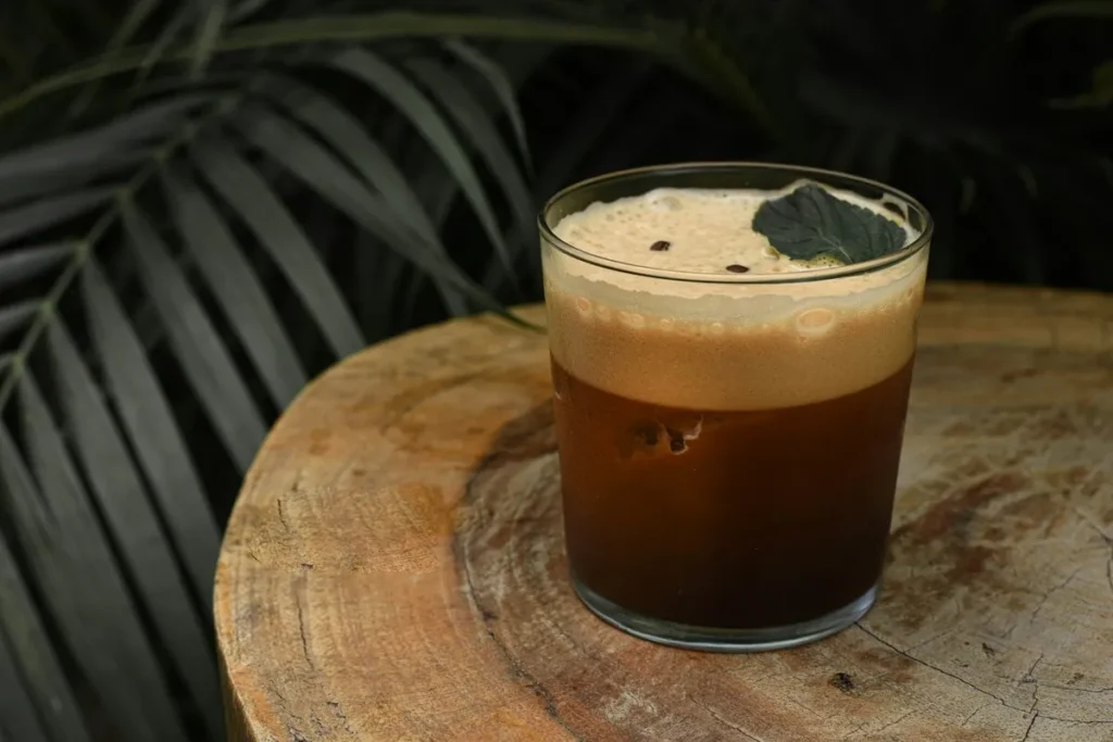 Um copo de carajillo, um coquetel espanhol feito com café, licor e gelo, decorado com grãos de café e uma folha de hortelã, sobre um tronco de madeira com folhagem escura ao fundo. A bebida escura com espuma clara representa o sabor intenso e a cultura do café.