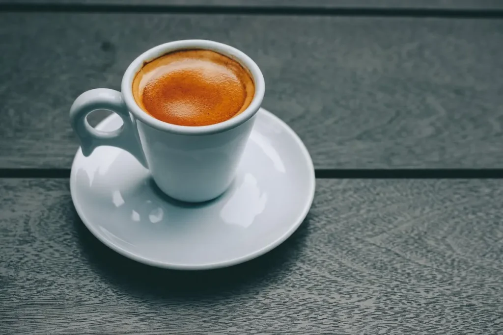 Uma pequena xícara de café expresso com espuma cremosa, sobre um pires branco, em uma mesa de madeira escura.