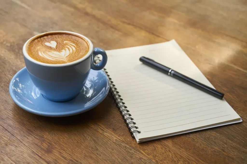Uma xícara de café com arte latte em forma de coração, acompanhada de um caderno e uma caneta sobre uma mesa de madeira.