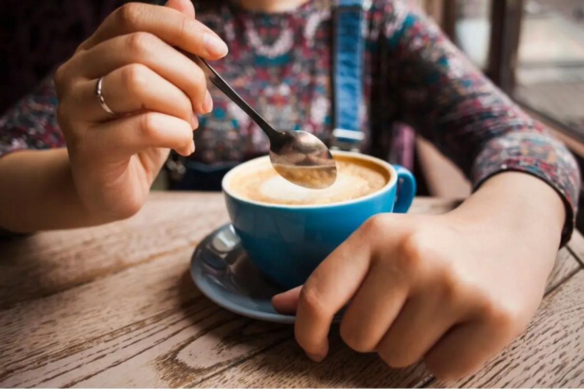 Pessoa segurando uma xícara de café com leite enquanto mexe a bebida com uma colher em uma mesa de madeira.