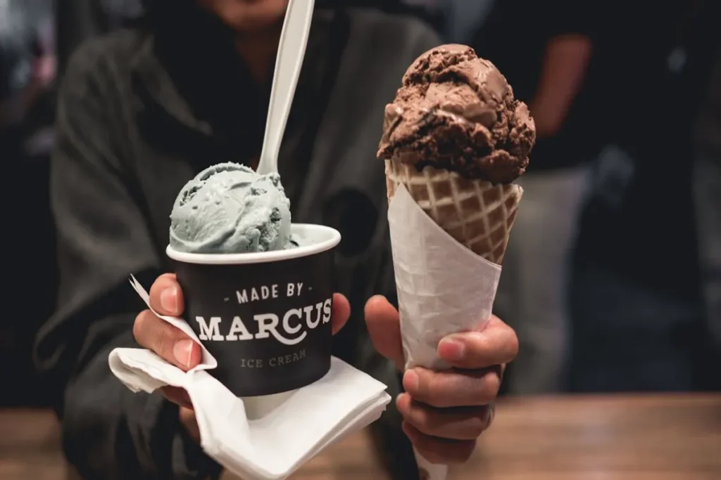 Duas mãos seguram um pote de sorvete de pistache ou menta e uma casquinha de sorvete de chocolate. A imagem simboliza a experiência de desfrutar de café e sobremesas.