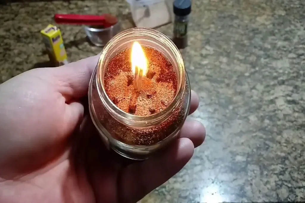 Mão segurando um pote de vidro com uma vela artesanal acesa, feita com pó de café moído. O pavio está em chamas, iluminando a superfície granulada do café, ilustrando a palavra-chave "queimar pó de café" para repelir mosquitos e perfumar o ambiente.