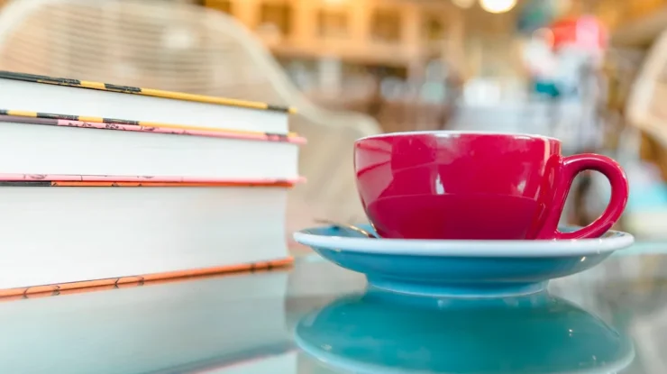 Xícara de café vermelha sobre um pires azul ao lado de uma pilha de livros em uma mesa de vidro, com fundo desfocado de uma livraria aconchegante