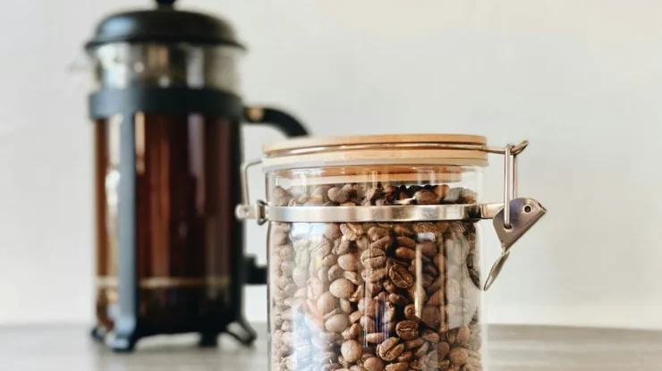 Pote de vidro com tampa de madeira contendo grãos de café torrados, posicionado em uma mesa com uma prensa francesa desfocada ao fundo.