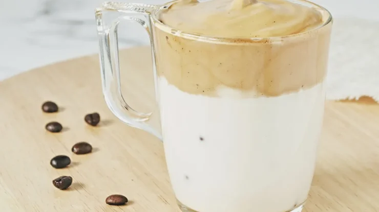 Xícara transparente com leite branco na base e espuma cremosa de café batido no topo, decorada sobre uma superfície de madeira com grãos de café ao redor.