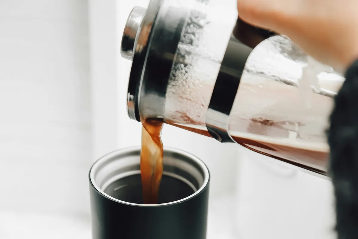 Café sendo servido de uma prensa francesa para uma caneca térmica preta, destacando o processo artesanal de preparo.