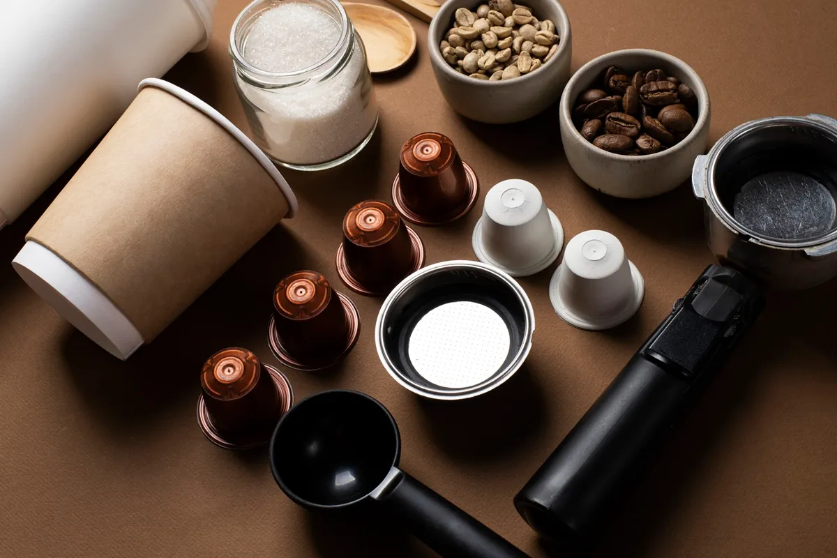 Utensílios e ingredientes para café, incluindo cápsulas, grãos crus e torrados, porta-filtro e copos descartáveis, organizados sobre uma superfície marrom.