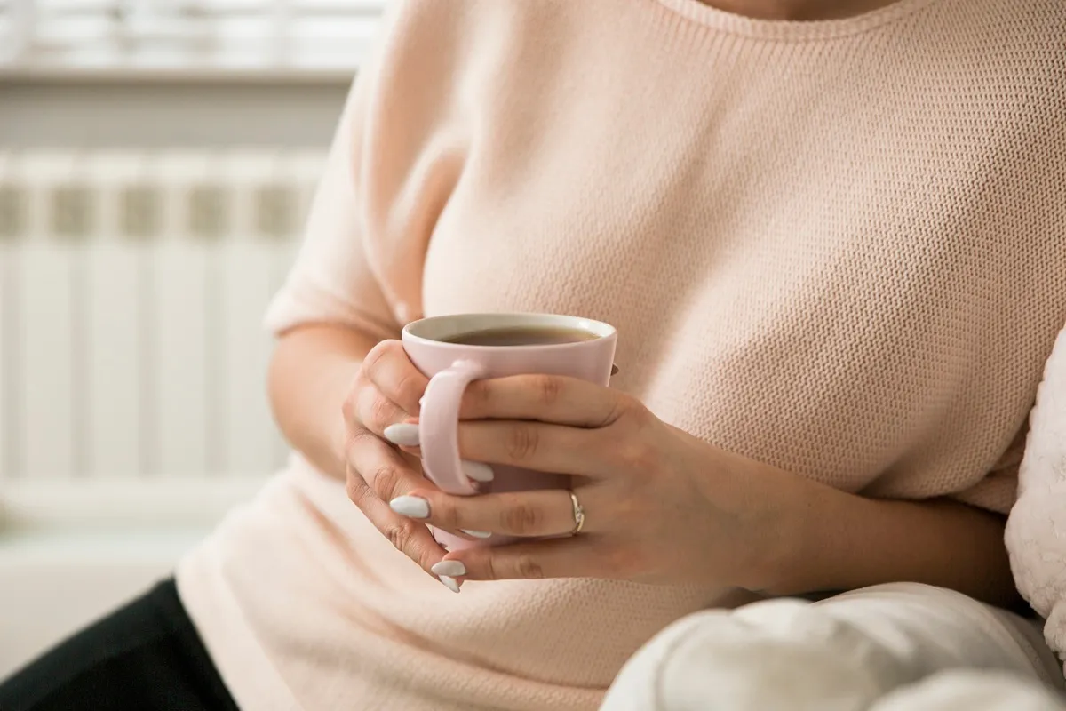 Pessoa segurando uma xícara rosa de café quente com as mãos, usando uma blusa bege e sentada confortavelmente em um ambiente aconchegante.