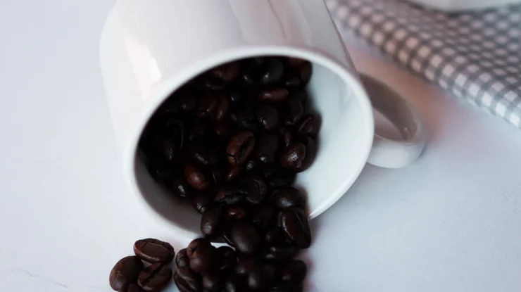 Grãos de café torrados espalhados de uma xícara branca virada sobre superfície clara, com pano quadriculado ao fundo.