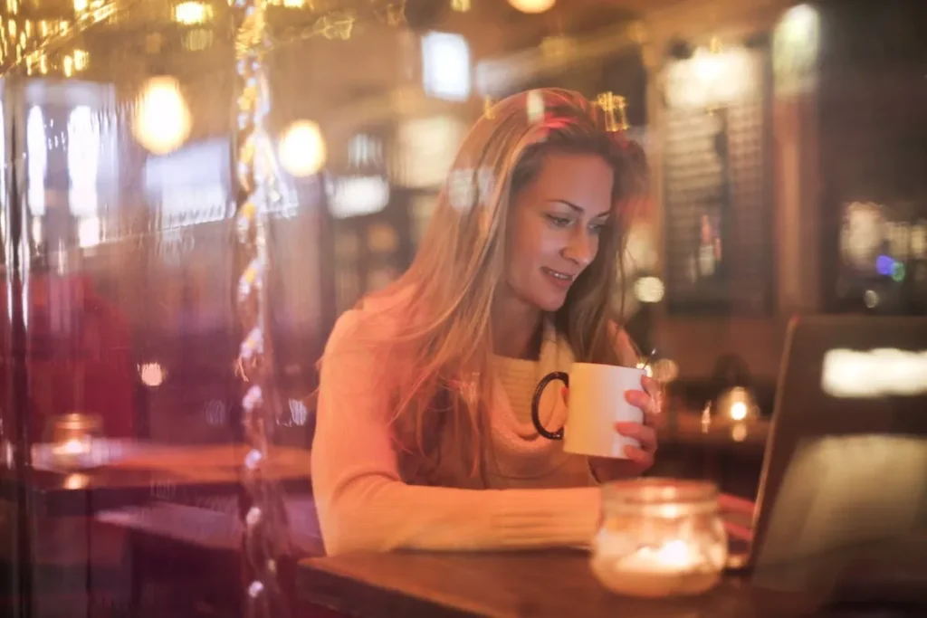 Mulher sorridente sentada em uma cafeteria, segurando uma caneca, enquanto olha para um notebook. A cena é vista através de uma janela embaçada e iluminada, sugerindo um ambiente aconchegante, ideal para refletir sobre o tema "pode tomar café com diarreia".