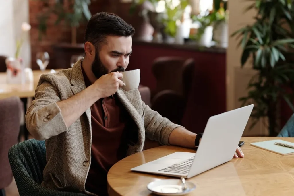 Homem barbudo, de terno e camisa, tomando café e olhando para seu notebook em um ambiente de escritório ou cafeteria. A cena ilustra a reflexão sobre o tema pode tomar café com diarreia.