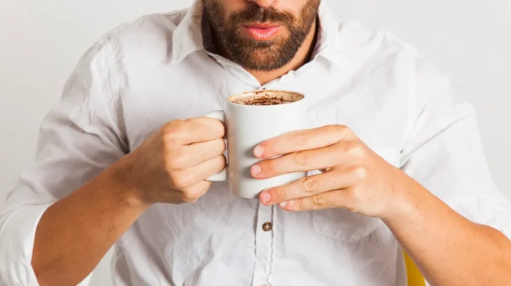 Homem de camisa branca soprando uma xícara de café quente enquanto a segura com as duas mãos.