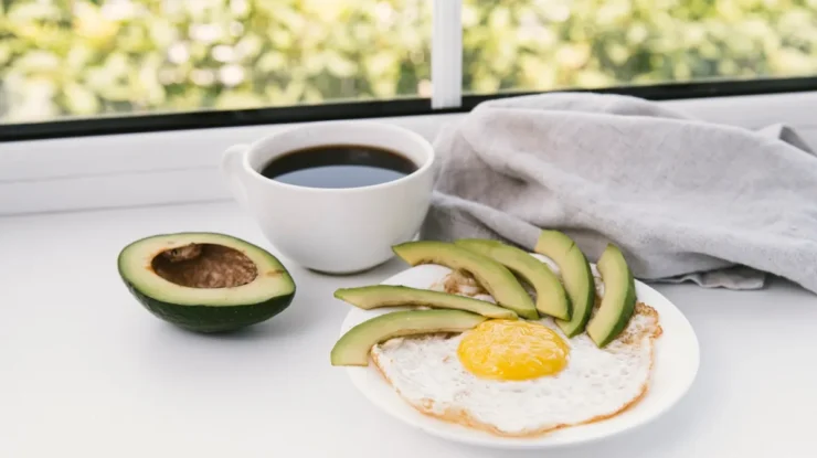 Prato com ovo frito e fatias de abacate, acompanhado de uma xícara de café e meio abacate ao lado, em uma janela iluminada.