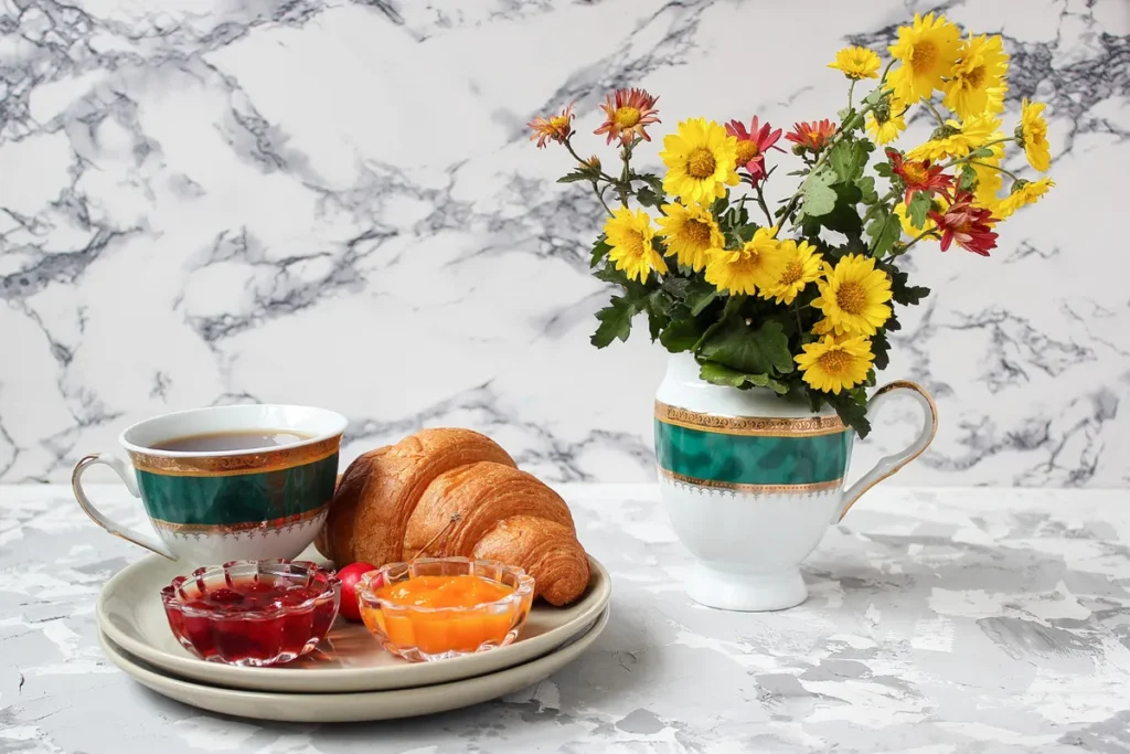 Um café da manhã de luxo com croissant fresco, geleias em tigelas de vidro, uma xícara de café e um arranjo de flores amarelas e vermelhas, sobre uma mesa de mármore.