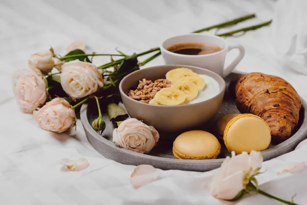 Bandeja com café da manhã de luxo na cama, com croissant, iogurte com banana, macarons e rosas cor de rosa, em um lençol branco.