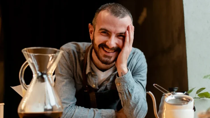 Barista sorridente com equipamento de preparo de café à frente, incluindo uma Chemex e chaleira, em um ambiente aconchegante.