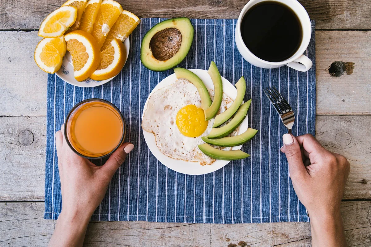 Prato com ovo frito, fatias de abacate, suco de laranja e café preto, acompanhado de fatias de laranja fresca, sobre uma toalha listrada.